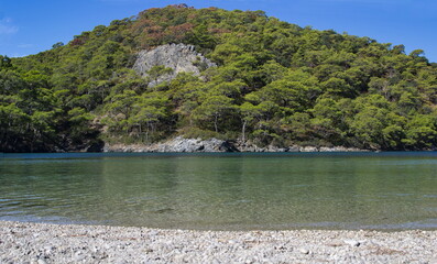 The clear water of the Blue Lagoon in Oludeniz and the beach with small pebbles and a green mountain in the background.