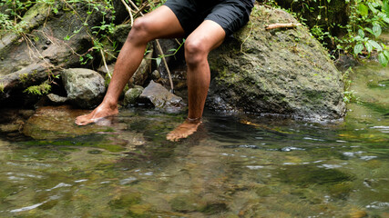 Barefoot in Clear Stream: Man’s Legs Resting on Wet Rocks Amid Green Forest