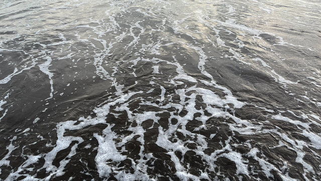 a gentle ocean wave washing over a dark sandy shore, creating foam patterns and textured water. natural beach scene with calm movement and simple composition - Powered by Adobe