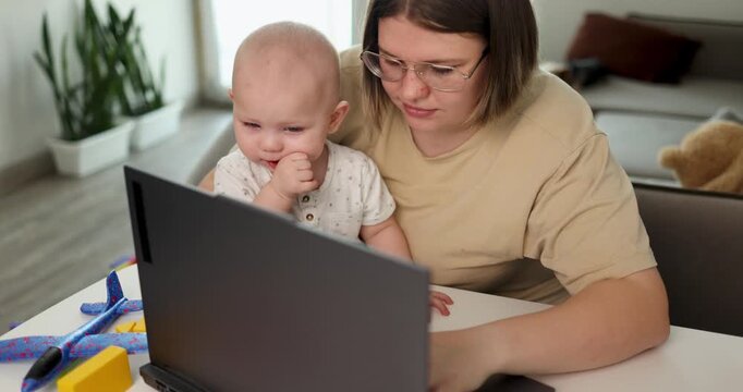 caucasian mother focused with baby and laptop, hands typing on keyboard, plants in background, concentrated