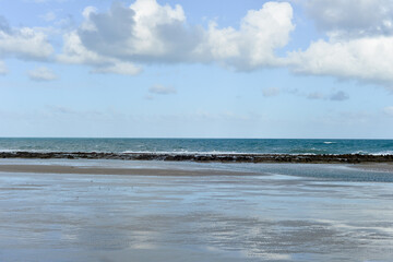 wet sandy beach at low tide with calm sea and rocky reef, tranquil coastal landscape under partly cloudy sky, peaceful reflective mood, horizontal orientation
