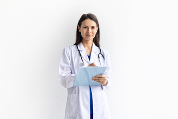 Smiling female doctor wearing white medical coat over blue uniform writing on clipboard on white background, confident professional healthcare worker