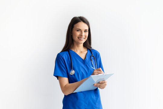 Smiling young female doctor in blue scrubs holding clipboard and pen on white studio background, confident medical professional posing for healthcare concept, copy space - Powered by Adobe