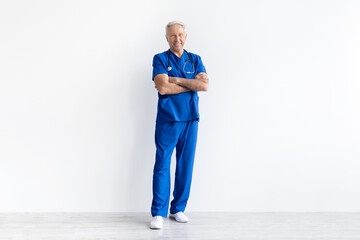 Senior male doctor in blue uniform standing with arms crossed on white studio background, confident experienced healthcare professional portrait