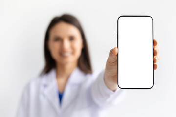 Smiling female doctor wearing white medical coat holding smartphone with blank screen toward camera on white background, healthcare technology and telemedicine concept