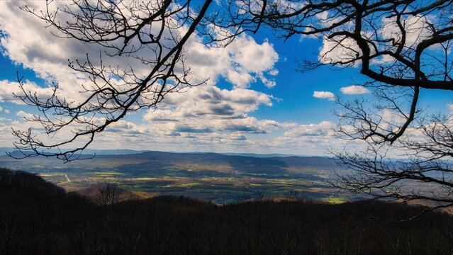 Bare branches frame a bright valley below the rolling Blue Ridge under a sky of scattered spring clouds.
