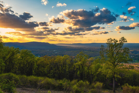Warm evening light spills across spring foliage and distant Blue Ridge ridges beneath dramatic clouds.