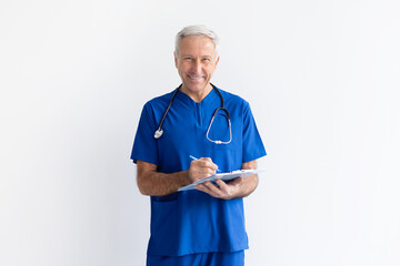 Smiling senior male doctor in blue scrubs writing on clipboard on white background, professional healthcare specialist recording medical notes