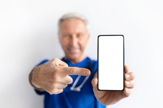 Smiling senior male doctor in blue uniform pointing at smartphone with blank screen on white background, healthcare technology and telemedicine concept