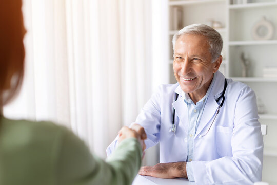 Smiling senior male doctor shaking hands with female patient in bright clinic, positive healthcare interaction and trust concept