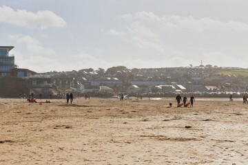 A sunny beach scene with people walking along the sand. Buildings are visible in the distance, creating a coastal landscape in Perranporth - Cornwall - UK