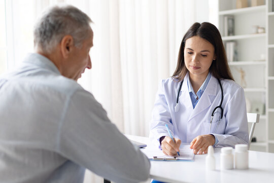 Female doctor in white coat writing medical notes during consultation with senior male patient in clinic, focused healthcare professional at work - Powered by Adobe