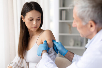 Senior male doctor applying adhesive bandage on female patients arm after vaccination in clinic, healthcare immunization and medical protection concept
