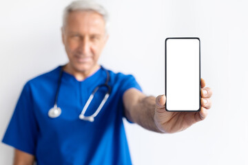 Smiling senior male doctor in blue medical uniform holding smartphone with blank screen toward camera, healthcare communication and digital technology concept