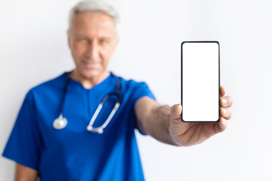 Smiling senior male doctor in blue medical uniform holding smartphone with blank screen toward camera, healthcare communication and digital technology concept