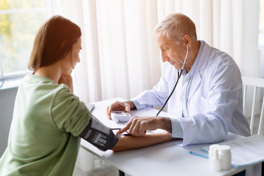 Senior male doctor measuring blood pressure of female patient in clinic using stethoscope, healthcare examination and medical checkup concept