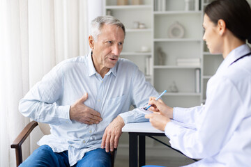 Senior male patient holding chest while talking to female doctor in white coat during medical consultation in clinic, healthcare and diagnosis concept