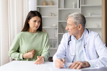 Fototapeta premium Pregnant woman feeling unwell, touching stomach and talking to senior male doctor in clinic office, maternity healthcare and pregnancy care concept