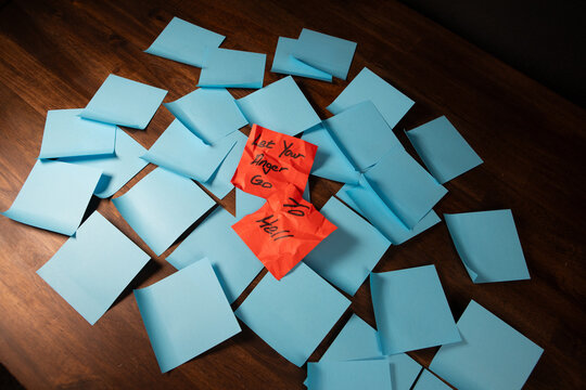 Crumpled red sticky notes on a pile of blue sticky notes a dark wood surface reading "Let Your Anger Go to Hell"