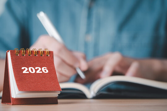 Showing a 2026 desk calendar with a person writing in a notebook representing planning organization future goals time management and preparation for the upcoming year in business and personal life