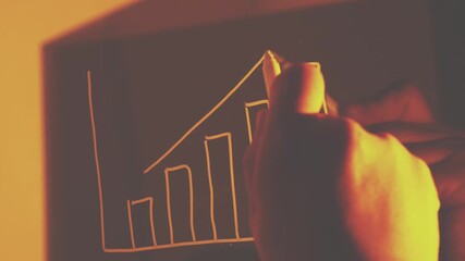 A close-up shot of a hand drawing an upward-trending bar and line graph on a dark surface, bathed in warm, orange light. - Powered by Adobe