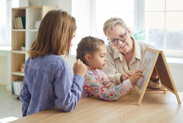 Smiling grandmother looking at her grandchildren playing with colorful geometric jigsaw, stacking puzzles into picture. Cheerful woman assisting kids with educational game during family leisure time.