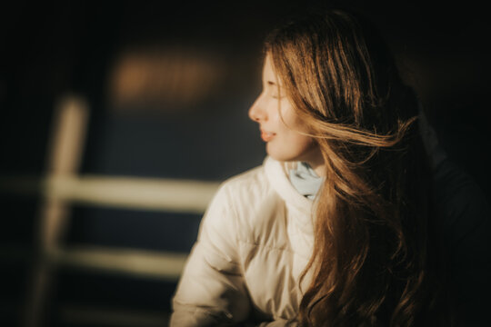 A serene portrait of a young woman with long hair, wearing a white winter jacket, looking away from the camera into the warm sunlight. Her hair catches the light beautifully, creating a soft, dreamy a