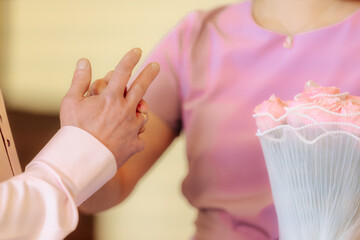 A tender moment showing a couple holding hands, both wearing wedding rings. Soft lighting and pastel clothing suggest a romantic or ceremonial occasion.