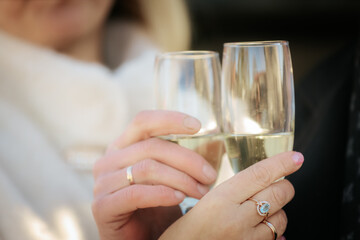 A romantic close-up of a couple clinking champagne glasses, both wearing wedding rings. The focus is on their hands and sparkling wine, suggesting celebration or engagement.