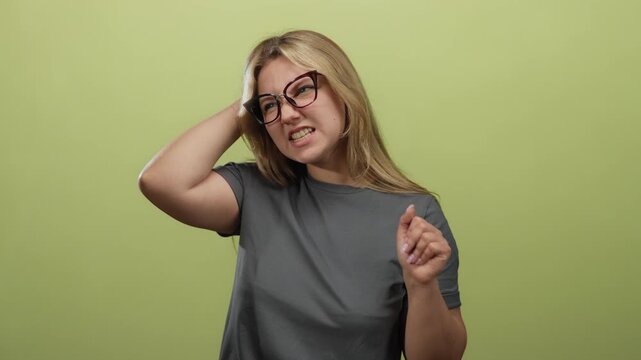 Woman gesturing with hand on head showing frustration against green background wearing glasses and casual grey shirt.