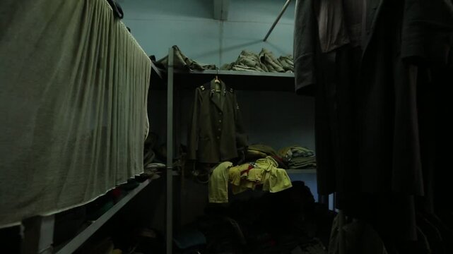 Slow zoom through dark military barracks storage room filled with uniforms hanging on racks, ending on a close-up display of many stacked, colorful officer's peaked caps.