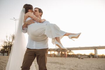 Romantic newlyweds on the beach at sunset, groom holding bride in white dress and veil, tender wedding moment outdoors
