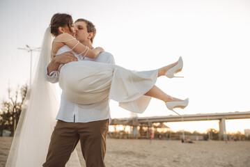 Romantic newlyweds on the beach at sunset, groom holding bride in white dress and veil, tender wedding moment outdoors