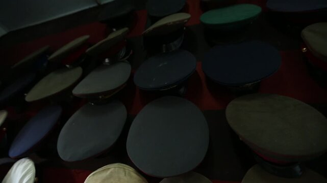 Slow zoom through dark military barracks storage room filled with uniforms hanging on racks, ending on a close-up display of many stacked, colorful officer's peaked caps.