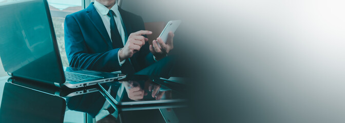 Businessman in blue suit working at desk using smartphone, laptop at the same time, highlighting multitasking, digital communication, corporate finance with clean blank space ideal for business copy.