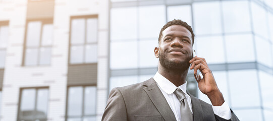 A businessman wearing a suit stands in front of a contemporary office building while talking on his phone. He looks confident and engaged, with bright sunlight illuminating the scene.