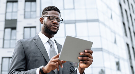 A professional man dressed in formal business attire is focused on a tablet while standing outside modern buildings on a sunny day. He appears engaged with the device.