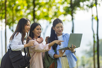 Group of Asian college students standing on green grass and discussing with laptop and books. Concept of teamwork, education, and outdoor learning in a natural environment.