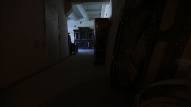 A long, dimly lit, and dusty corridor in an abandoned building with crumbling walls, open doorways, and debris covering the floor. Daylight shines faintly from the far end