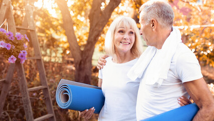 A smiling older couple carries yoga mats in a vibrant garden filled with autumn colors. They share a joyful moment as the sun sets, reflecting their active lifestyle.