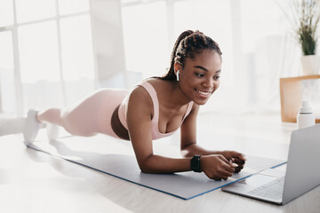 A sporty millennial black woman performs an elbow plank exercise on a mat in her home gym. She is...