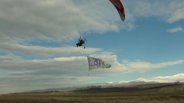 A motorized paraglider (paramotor/powered parachute) flies across a cloudy sky, towing a white advertising banner. Recreation, extreme sport, and aerial advertising activity.