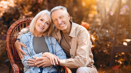 A happy couple relaxes together in a cozy wicker chair during a sunny autumn afternoon. They sit in a garden filled with vibrant autumn leaves, enjoying each other's company.