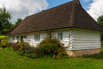 A white log cottage with a dark shingle roof stands amid a lush garden full of colorful flowers. The vibrant greenery and wooden details highlight rural craftsmanship, warm seasonal atmosphere, and au
