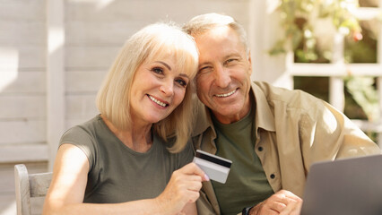 A joyful couple sits together at a table, smiling while holding a credit card. They are engaged in online shopping using a laptop, surrounded by a bright and inviting home environment.