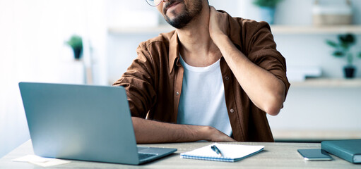 A young man sits at a desk, looking at his laptop. He shows signs of discomfort, rubbing his neck while taking notes. The workspace is well-lit with plants in the background.