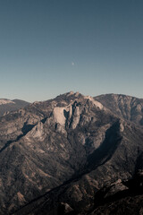 Fototapeta premium A rugged mountain landscape viewed from Moro Rock, with dramatic granite peaks and a faint moon beneath a clear blue sky.