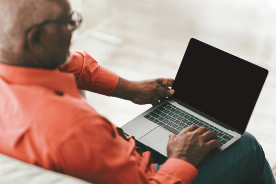 An elderly man is seated on a couch, focused on his laptop in a bright indoor space. He wears a red shirt and appears engaged with technology, highlighting modern connectivity.