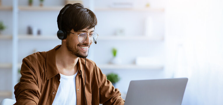A young man sits at a desk wearing a headset and smiling while he engages in an online meeting on his laptop. The bright room is filled with plants and soft light.