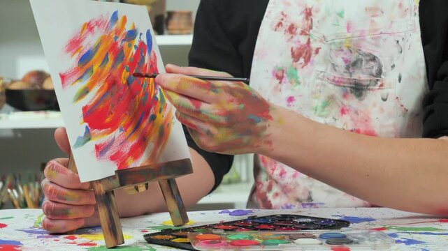 An artist in a white stained apron applies brushstrokes to a future painting.